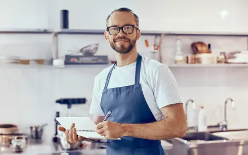 Ein Koch steht in einer professionellen Küche und notiert etwas auf einem Schreibblock, möglicherweise Rezepte, Bestellungen oder Vorbereitungspläne.