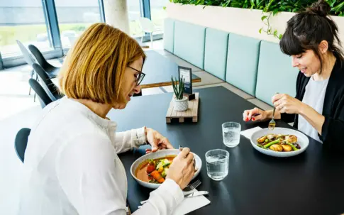 Zwei Frauen sitzen gemeinsam an einem Tisch in einer Kantine und unterhalten sich bei einer Mahlzeit.