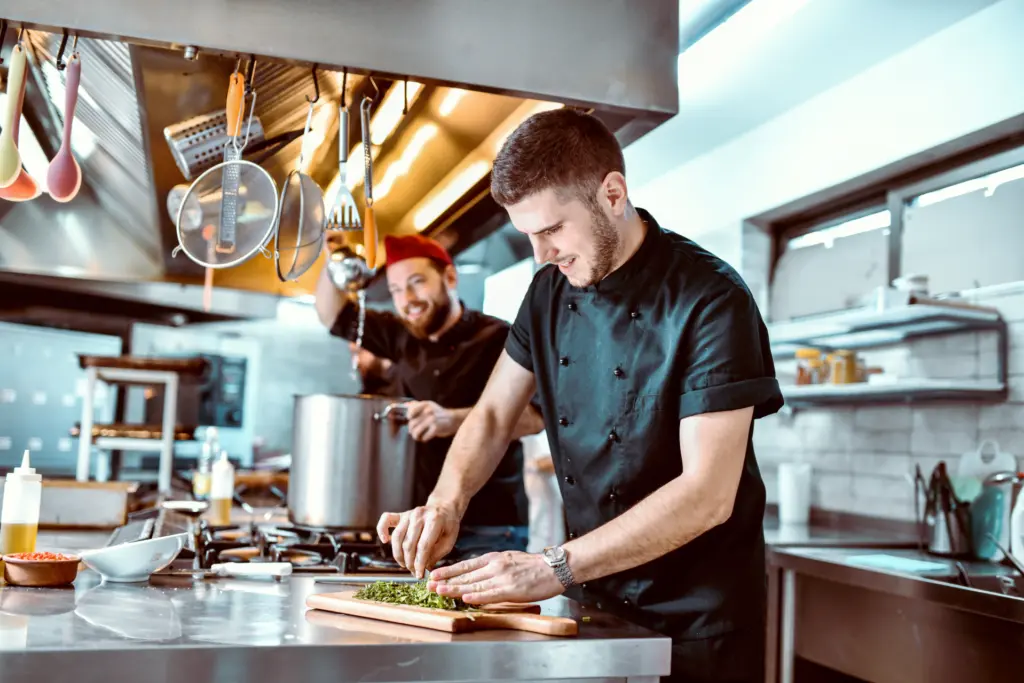 Zwei Köche stehen in einer professionellen Küche und bereiten Speisen zu, umgeben von Küchengeräten und frischen Zutaten.
