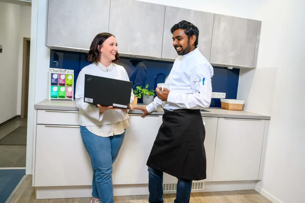 Manou und ein Kollege unterhalten sich in der Kaffeeküche, umgeben von Kaffeemaschine und Küchenutensilien.