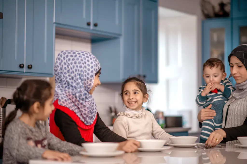 Familie sitzt in einer Küche zusammen, zwei Frauen mit Kopftuch, zwei Kinder und ein Baby.