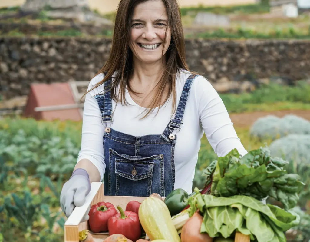 Frau mit einer Holzkiste voller frisch geerntetem Gemüse auf einem Feld.