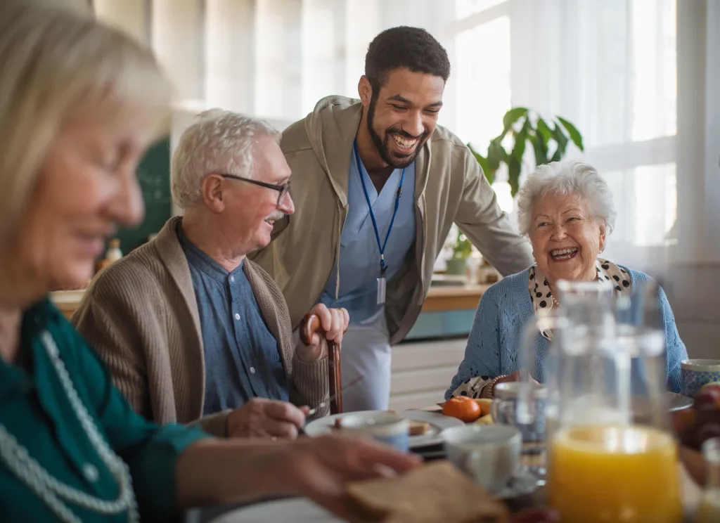 Fröhliche Senior:innengruppe beim Frühstück. Im Hintergrund steht eine Servicekraft.