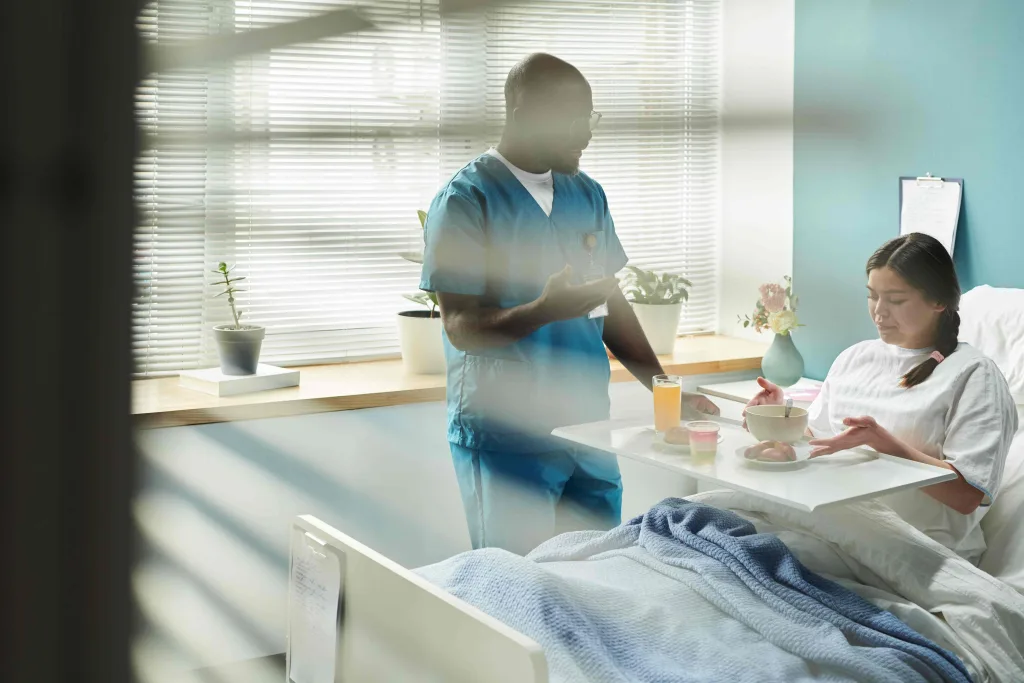 Ein junger Pfleger serviert einer Frau im Krankenhausbett das Frühstück