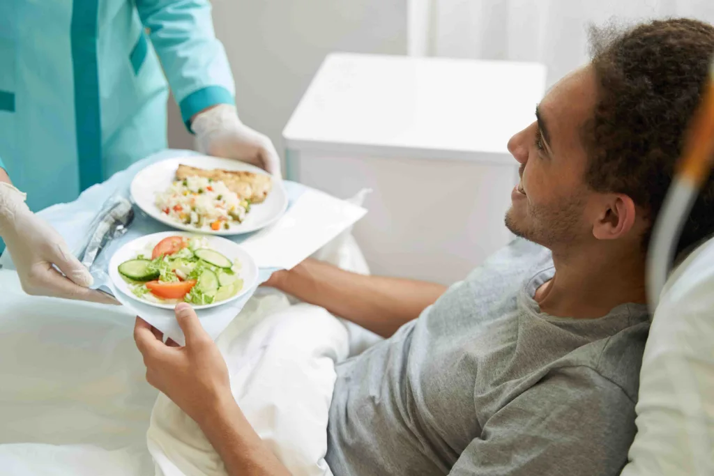 Ein junger Mann im Krankenbett bekommt einen gesunden Salat und Rohkost gereicht.