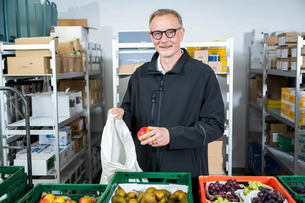 Mann hält Apfel und Stoffbeutel zwischen Kisten mit Obst in einem Lagerraum.
