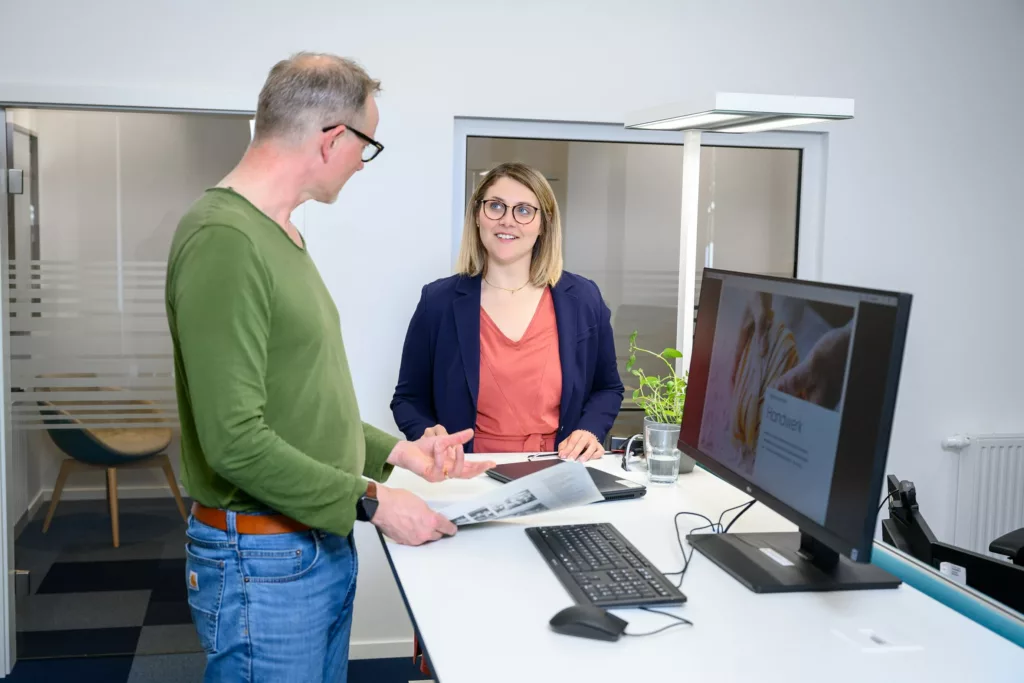 Alexander Gundlach spricht mit einem Kollegen an einem Schreibtisch mit Computer.