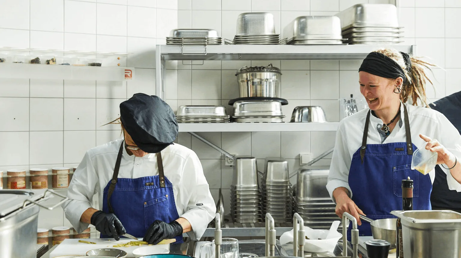 Zwei Frauen bereiten zusammen eine Mahlzeit in einer großzügigen, professionell ausgestatteten Küche zu – umgeben von Kochutensilien, Töpfen und frischen Zutaten.