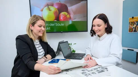 Zwei Frauen sitzen an einem Tisch mit Laptop und Unterlagen und besprechen ein Projekt.