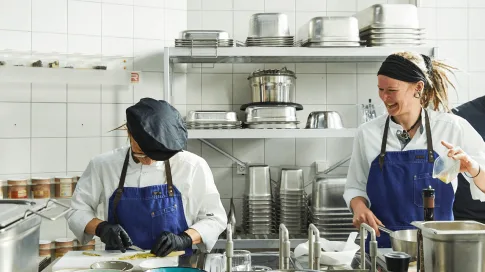 Zwei Frauen bereiten zusammen eine Mahlzeit in einer großzügigen, professionell ausgestatteten Küche zu – umgeben von Kochutensilien, Töpfen und frischen Zutaten.