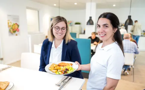 Eine Servicekraft übergibt einer Frau an einem Tisch einen Teller mit Essen.